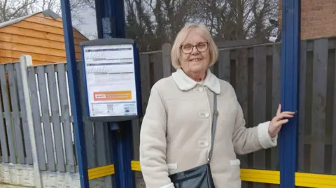 Glynis Chapman A woman with blonde hair in a bob and glasses is wearing a cream jacket with a black crossbody bag. She is standing at a bus stop and smiling.