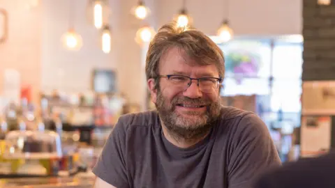 Nick Smith Nick Smith, who runs the Ludoquist board game cafe, sitting in his cafe, smiling at someone off camera