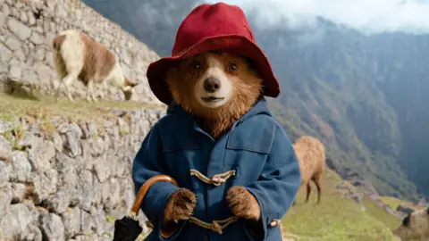 A still of Paddington Bear from the film Paddington in Peru wearing his trademark red hat and duffelcoat. Mountains and goats are in the background.