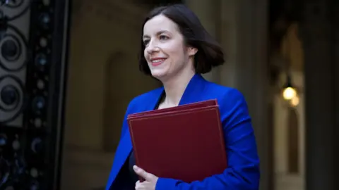 Bridget Phillipson smiling, wearing a blue suit and carrying a dark red folder