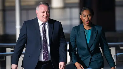 Getty Images Conservative Senedd leader Darren Millar walking alongside Kemi Badenoch in front of the Senedd building on a sunny day. They are talking to each other and looking ahead.