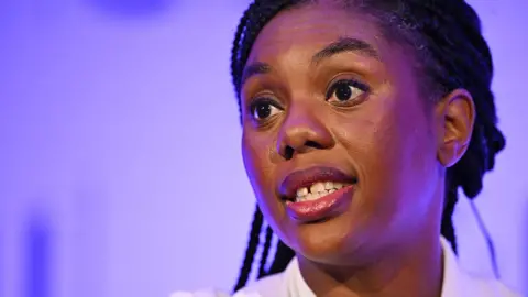 Close up headshot of Kemi Badenoch speaking against a purple background in London in November