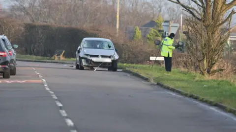Phil Taylor A car with a smashed windscreen and damage to its front is pulled up on the side of the road. A police officer in a high-vis jacket stands on the grass to the right
