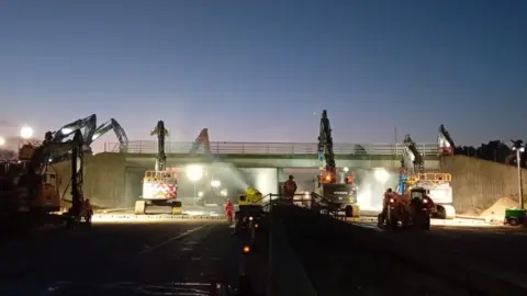 Various plant vehicles on a road demolishing a bridge above. The sun is setting behind the bridge