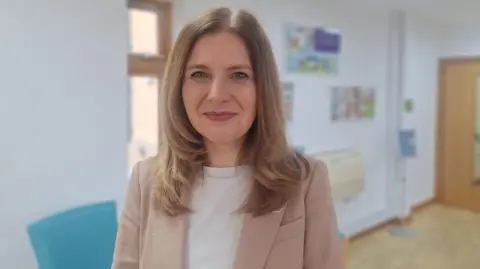 A woman stood in a corridor in a hospice. The background is blurred and she is in focus. She has shoulder-length blonde/brown hair and smiles at the camera. She has pink lipstick on and wears a beige jacket over a white t-shirt. It is a head and shoulders shot. 