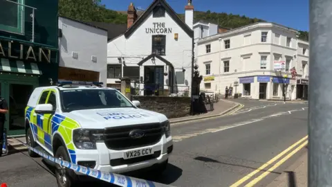 BBC A police car and blue and white striped police tape in the foreground with an empty street behind and on the other side of the road a number of white buildings, one with the words The Unicorn on it