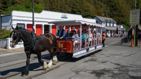 A dark brown horse with a white face and three white feet pulling a red and white open-sided tram out of the terminus in Douglas with the tram sheds behind. The tram is full of passengers and the driver is wearing a blue shirt and dark waistcoat.