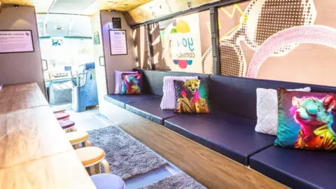 Langlois Photography The image show the interior of the bus. It has a large seating area with a long bench with dark cushions and decorative pillows. There is also a desk area with lots of little stools underneath.