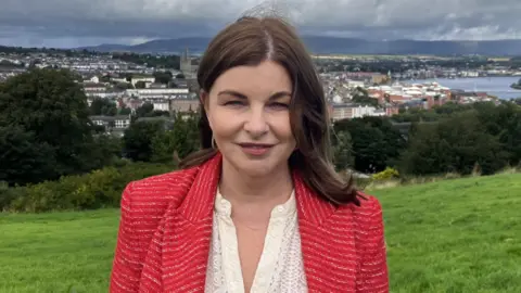 Shows a woman in a red blazer with a cream open neck top standing in a field with trees in the background and a river and buildings beyond that.