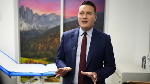 Wes Streeting is standing in front of a medical bed and a mural talking. He has dark brown hair gelled to stand up and blue eyes. He is wearing a blue jacket over a pale blue tie and burgundy tie. His hands are outstretched in front of him to emphasise what he is saying. The mural shows mountains and meadows under a red sky.