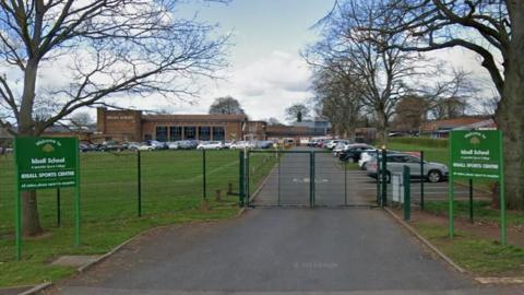 The front gates of Idsall School, with a road leading to the building surrounded by cars