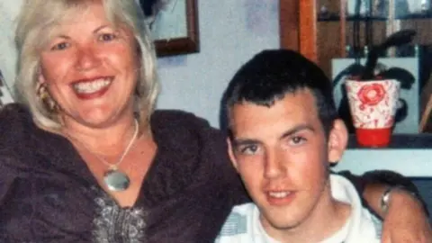 PA Media Melanie Leahy and her son Matthew sit side by side in the living room. The picture is slightly grainy. Matthew has dark brown, short cropped hair and is looking at the camera. His mother has a dark purple cardigan on and is beaming at the camera, so you can see her teeth, she is wearing a large necklace. There is a pot plant in the background.