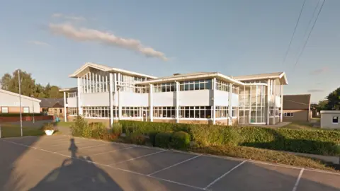 Google Maps Wymondham College, in Norwich: a white two-storey building, with large glass-paneled windows running continuously on each floor. In front of the building there is a green hedge that separates it from the parking garage. 