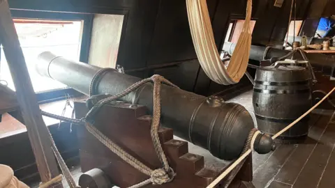 A canon sits under deck on the replica Galeon Andalucia. It sits beside a barrel, and is held in place by rope. It is pointing out a window