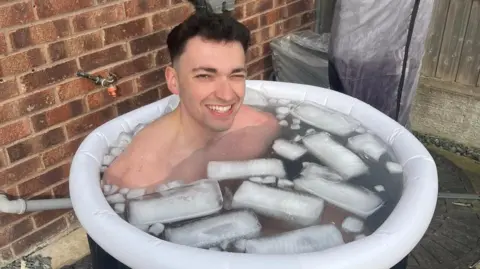 Nicola Byatt A topless man in an inflatable ice bucket, full of water and ice. He is smiling