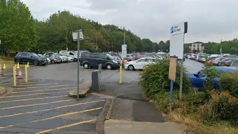 A car park with four long rows of parked cars. There are tall bushy trees in the background and to the left. There is a white parking sign to the right-hand side of the car park entrance. Also at the entrance, diagonal yellow line road markings lead to the parking area. 