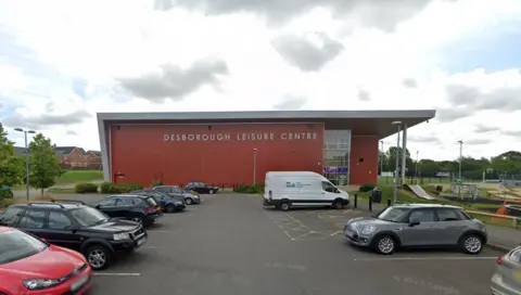 Google Brick building labelled "Desborough Leisure Centre" with a car park in front. Tree-lined paths are visible along with a skate ramp