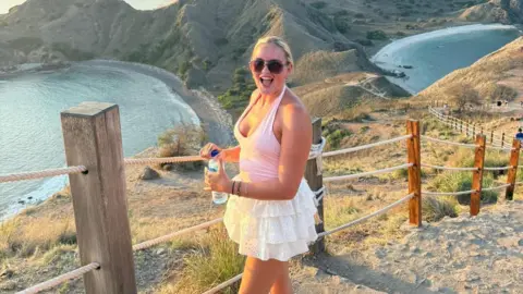 Regan Sparks A woman with blonde hair tied back looks delighted at the camera. She is wearing a pink strappy top and a ruffled white skirt. A sun-dappled bay is behind her.