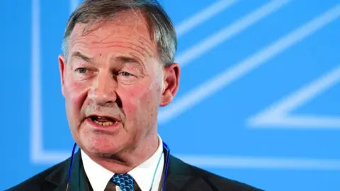 PA Media A close-up of Rupert Lowe. He has short grey hair and is looking to his right while giving a speech. He is wearing a grey suit jacket over a white shirt and blue tie. Behind him there is a blue coloured screen. 