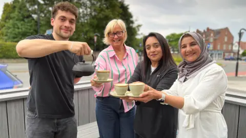 A young man with short, curly hair and a goatee beard poses with three ladies. He is posing pouring out tea from a vintage Middle Eastern style teapot while the ladies hold out their cups and saucers. They are all standing inside an enclosure with wooden benches and behind them is a busy street scene with a road, cars and buildings visible.