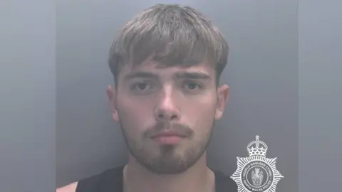 A mugshot picture of a man with brown hair and a short beard. He is wearing a black vest