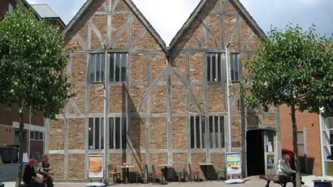 BBC / Josie Campbell Outside view of Pescod Hall which is a two-storey building with grey wooden frames and red bricks. There are two pitched roofs and small vertical windows on both floors. Chairs and tables from a neighbouring sandwich shop are at the front of the building.