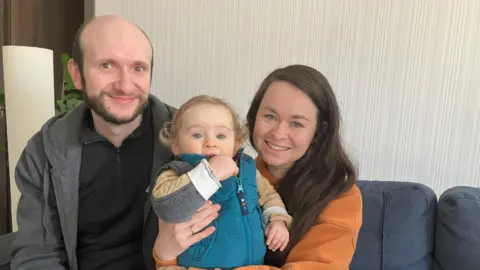 Ihor, in a grey top and black shirt, sits with Yulia, in an orange top, with their baby son Markiian between them dressed in a turquoise sleeveless top.