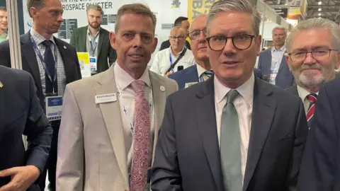 Farnborough Aerospace Consortium Keir Starmer stands in front of a group of men in suits on one of the stands in an exhibition hall at the air show.