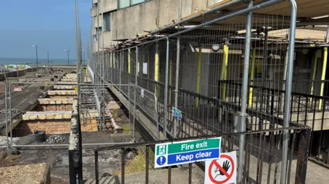 Metal fencing, scaffolding and temporary supports surround repair works outside a seafront tower block. Excavated sections of ground run alongside the building, with the sea visible in the distance and a sign reading "Fire exit Keep clear" attached to a gate.
