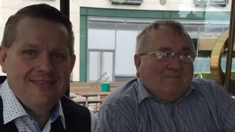Two men looking and smiling at the camera. One wears a black waistcoast and blue spotty shirt with short black hair. Another wears glasses and blue striped shirt with grey hair. 
