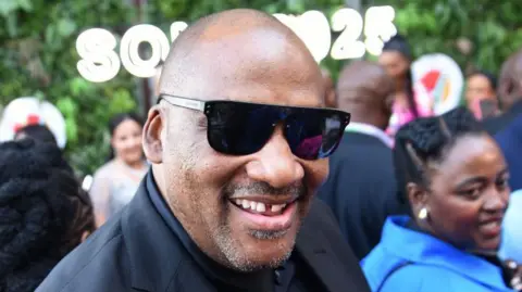 Gallo Images via Getty Images A close-up of Gayton McKenzie, who is wearing dark glasses and smiling at the camera - a gap in his teeth is visible.