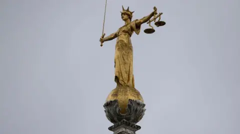  A statue of lady justice holding the scales of justice. The gold statue is on top of a stone pillar and curved holder. 