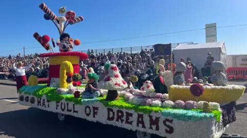 A flower display at the Battle of the Flowers event in 2024. The display says on the side 'Hat full of dreams' and there is a red and yellow hat on the left, mushrooms and candy canes made out of flowers.