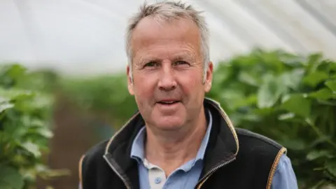 a man with grey hair wearing a blue shirt and gilet. he is standing in a polytunnel with plants growing behind him.