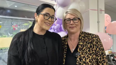 A photo of two women smiling looking into the camera. One woman has short blonde hair and the other has long black hair. Both are slightly smiling and looking into the camera. One has a leopard print jacket on and the other is wearing all black. 