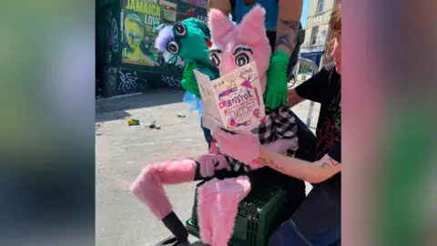 Bean Pig Puppets A pink puppet with pointy ears and a green, google-eyed puppet read the programme of the Bristol Experimental Puppet Festival. The puppeteers' arms are visible. 