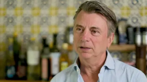 Michael Moszynski is wearing a subtly blue striped shirt in the background is an interior with different coloured bottles on a shelf and bright wallpaper.