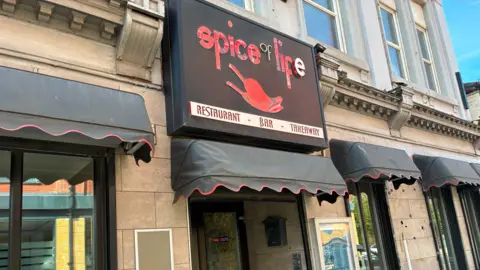 Exterior view of the Spice of Life restaurant. It has the name of the business detailed in red on a black sign above the door. There is a menu board to the side of the entrance.