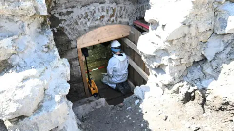 A member of the forensic excavation team working in the subterranean vaulted structure. The person is wearing a white overall suit.