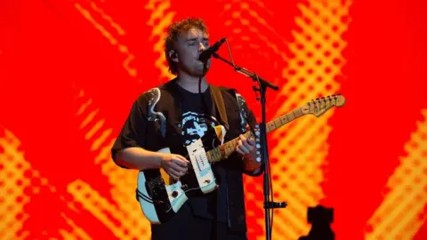 Getty Images A face on photo of a singer holding a guitar. The background is red from a projector. He is wearing a black top and has curly short hair.