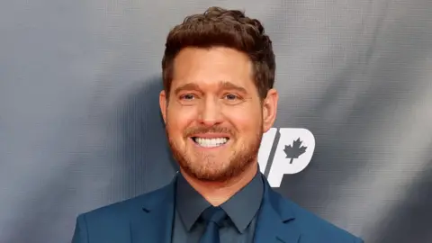 Michael Bublé wearing navy suit, shirt and tie. He has a wide smile on his face. 