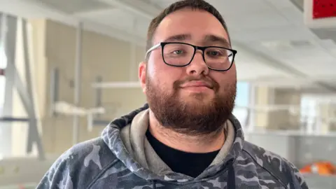 A man in his 20s with short dark hair and a beard, stands looking at the camera and smiling. He is wearing black framed glasses, a grey camouflage hoodie with a black T-shirt on underneath. He is stood in a room with hospital apparatus behind him.