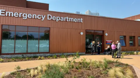 External view of the entrance to Arrowe Park Hospital's new Emergency care department. Patients and staff are see talking outside the front door. It is a sunny day.