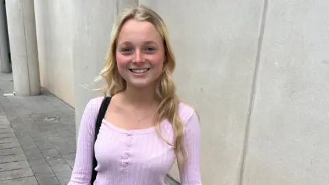 A young woman with long curly blonde hair. She is wearing a pale pink long sleeve top and has a black bag strap on her shoulder. She is smiling and looking at the camera.