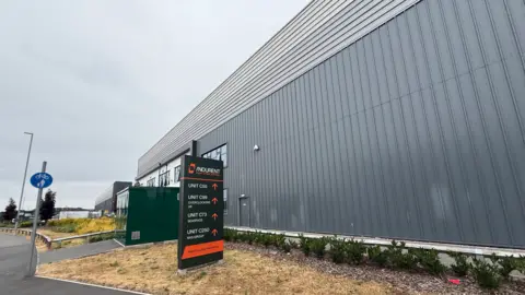 BBC The outside of a large grey facility at a site featuring industrial and logistics space. A mainly black sign outside the facility includes the word Indurent, as well as Unit C55 next to an arrow pointing the way to that. 