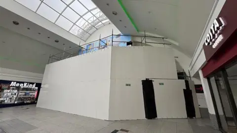 White hoarding in in place at the end of a shopping centre with a curved glass roof and a store either side of it, one called MenKind and one a Costa Coffee