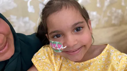 Saffanah smiling into the camera. She has dark hair died back and is wearing a yellow top with white flowers on it. She has a tube going into her nose and colourful tape on her face.