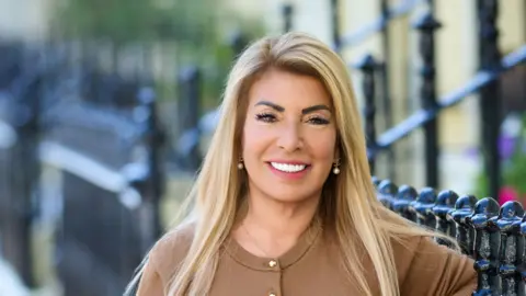 Jo Hemmings Head and shoulders of a woman with long straight hair in a smart sweater standing outside in front of a black metal railing.