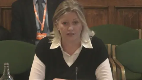 PA Media A woman with shoulder length blonde hair mid speech wearing a white shirt with a black vest over the top.