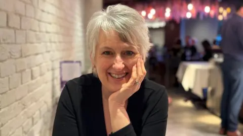 Erica May A woman with short grey hair smiling sat at a table with her chin resting on her hand.
